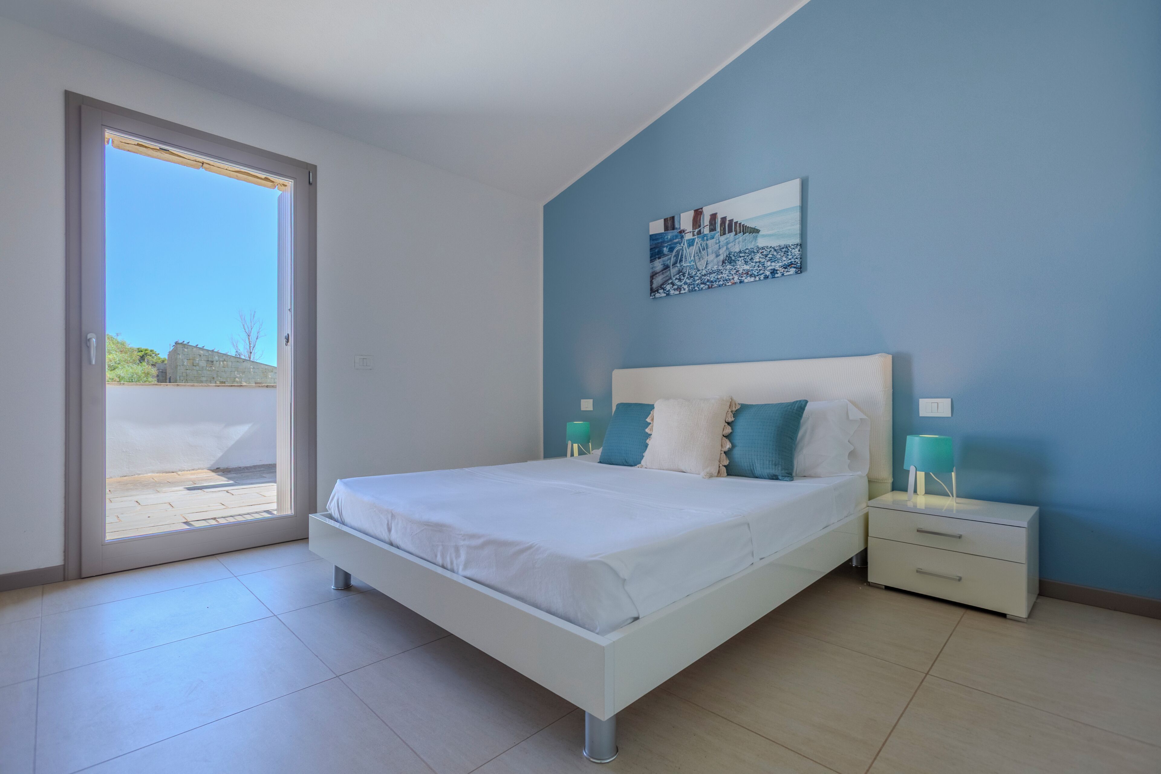 A bedroom with a bed, nightstands, a painting, and a view through a sliding door.