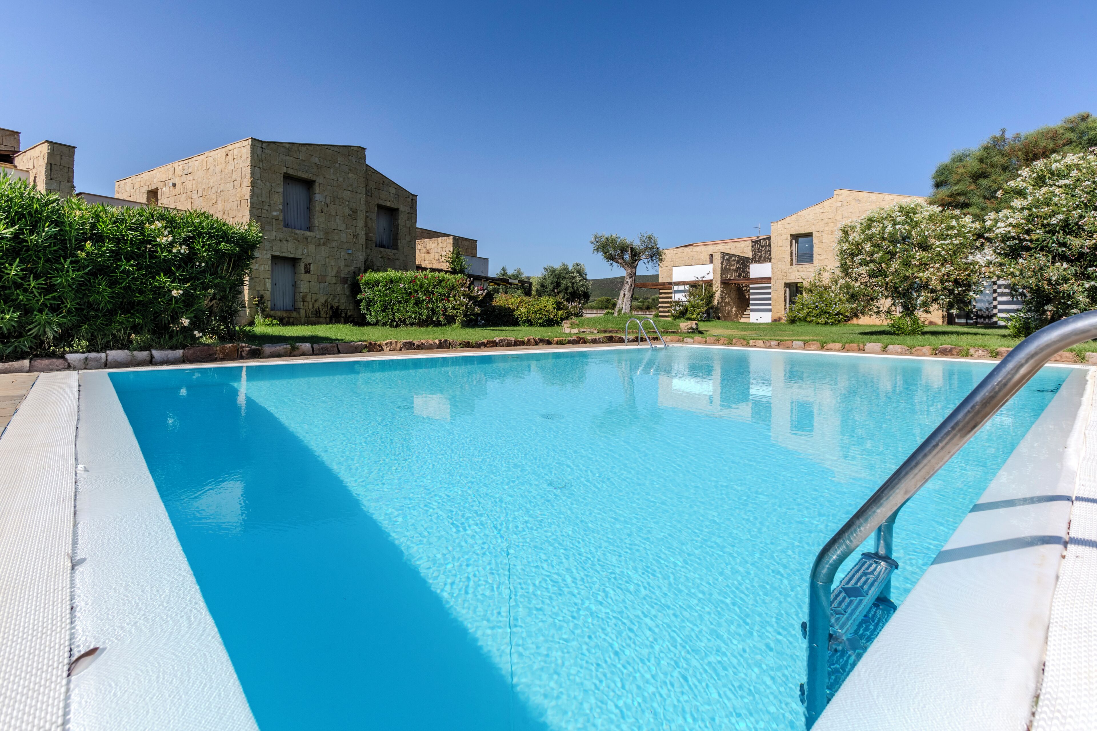 A clear blue swimming pool with a metal ladder, surrounded by stone buildings and greenery.