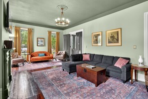 A spacious living room with a large sectional sofa, a coffee table, and a patterned rug. There are two doors leading to other rooms, and a chandelier hangs from the ceiling. - FLX Victorian, Historical Charm + Modern Comfort, Perfect Location to Everything (Ovid)