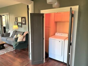 A laundry room with a washing machine and a door leading to a living room with a couch and a lamp. - FLX Victorian, Historical Charm + Modern Comfort, Perfect Location to Everything (Ovid)