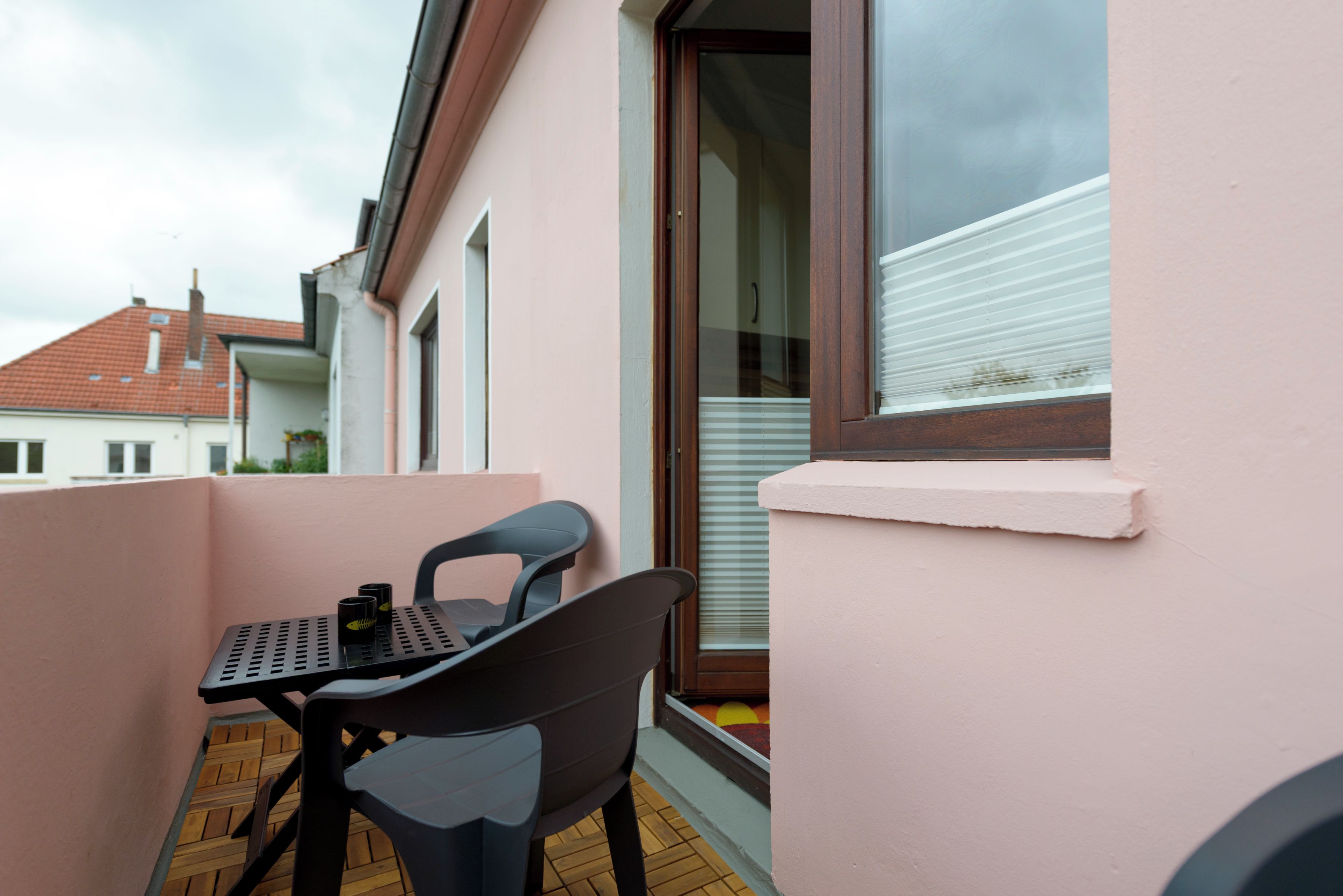 Uma varanda com mesa e cadeiras, uma janela com persianas e vista para edifícios vizinhos.
