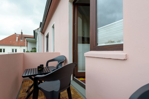 A balcony with a table and chairs, a window with blinds, and a view of neighboring buildings.
