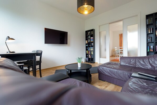 A modern living room with a sofa, a desk, a chair, a small table, a bookshelf, and a flat-screen TV mounted on the wall.