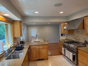 A modern kitchen with granite countertops, wooden cabinets, a stainless steel stove, and a dining area with a table and chairs. - Spacious 5-bedroom house in Falmouth within walking distance to Beach and Town (Falmouth)