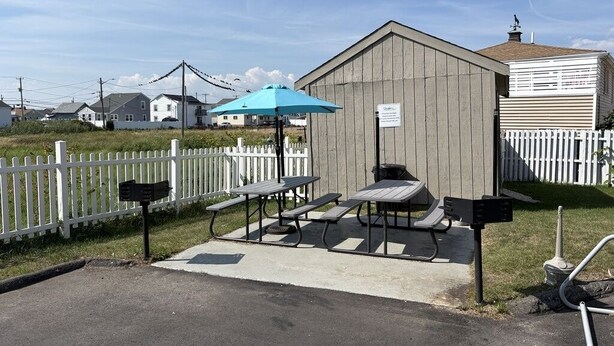 A picnic area with tables and an umbrella, a wooden building, and a white picket fence.