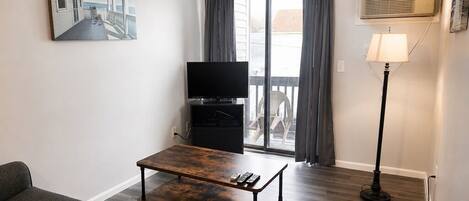 A living room with a sofa, a coffee table, a flat-screen TV, a balcony with curtains, and a standing lamp.