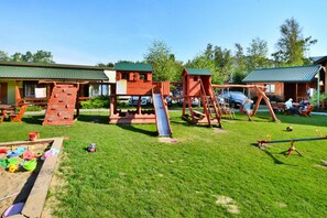 Ein Spielplatz mit Rutsche und Klettergerüsten, umgeben von einer Rasenfläche und Holzbauten.