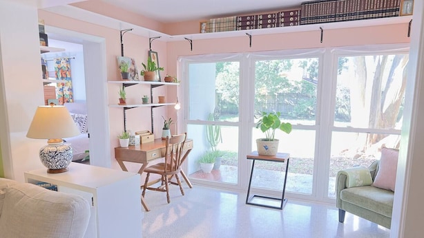 Una habitación acogedora con escritorio, silla y sofá verde. Hay estantes con libros y plantas, además de una ventana con vistas a árboles.