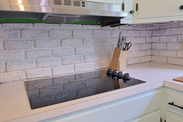 Une cuisine avec un mur de briques blanches derrière l’évier, une plaque de cuisson noire et un porte-ustensiles en bois contenant des ciseaux.