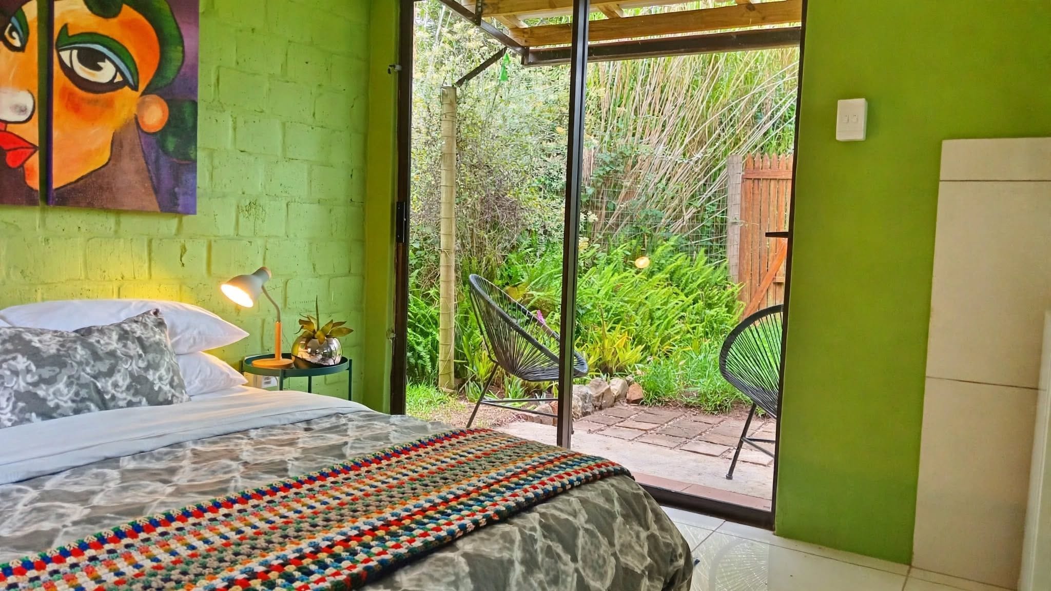 Een slaapkamer met een bed, een nachtkastje, een lamp, een kleurrijk vloerkleed en uitzicht op een tuin door een schuifdeur.