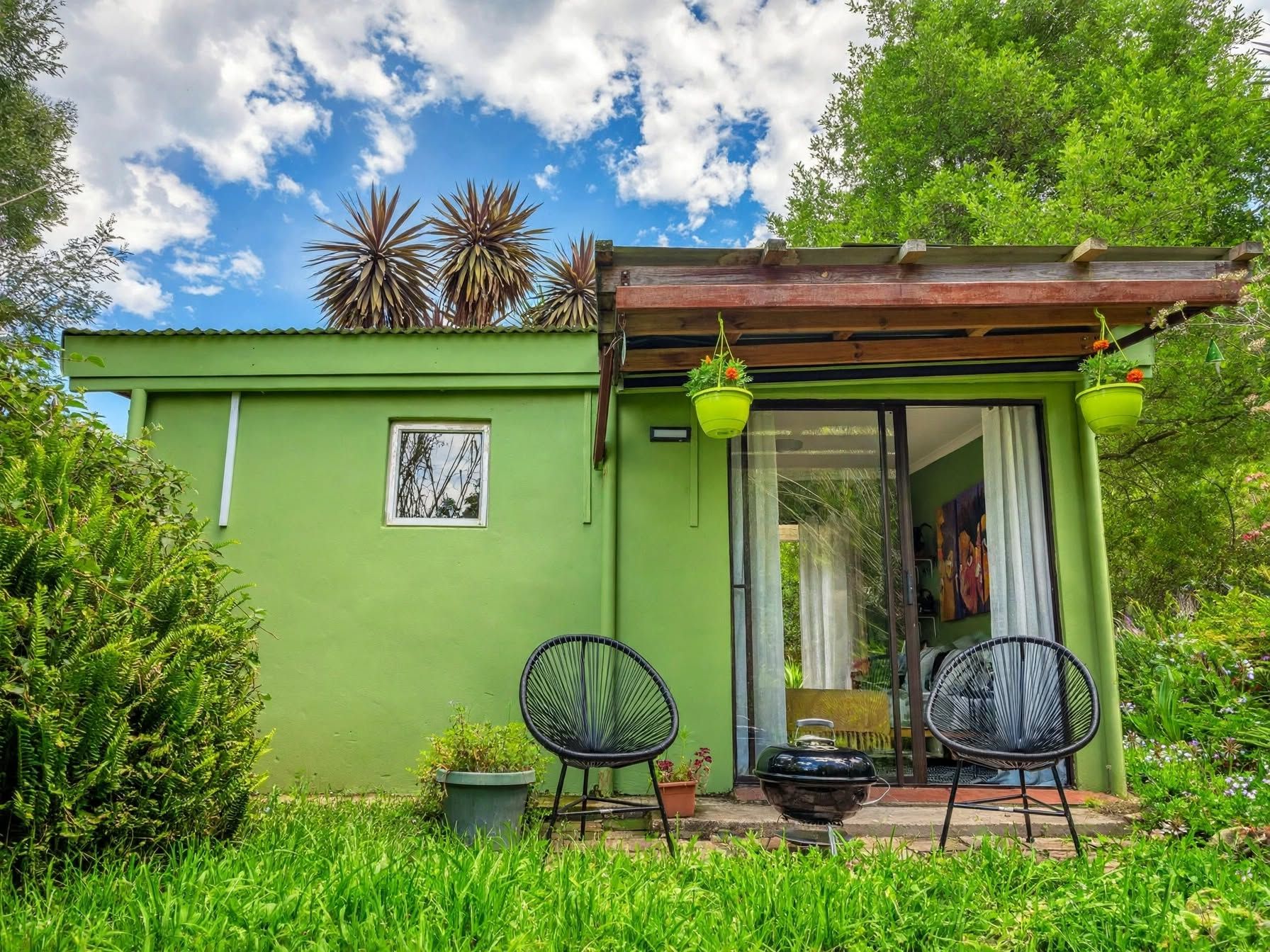 A green house with a sliding glass door, two chairs, a barbecue grill, and hanging planters.