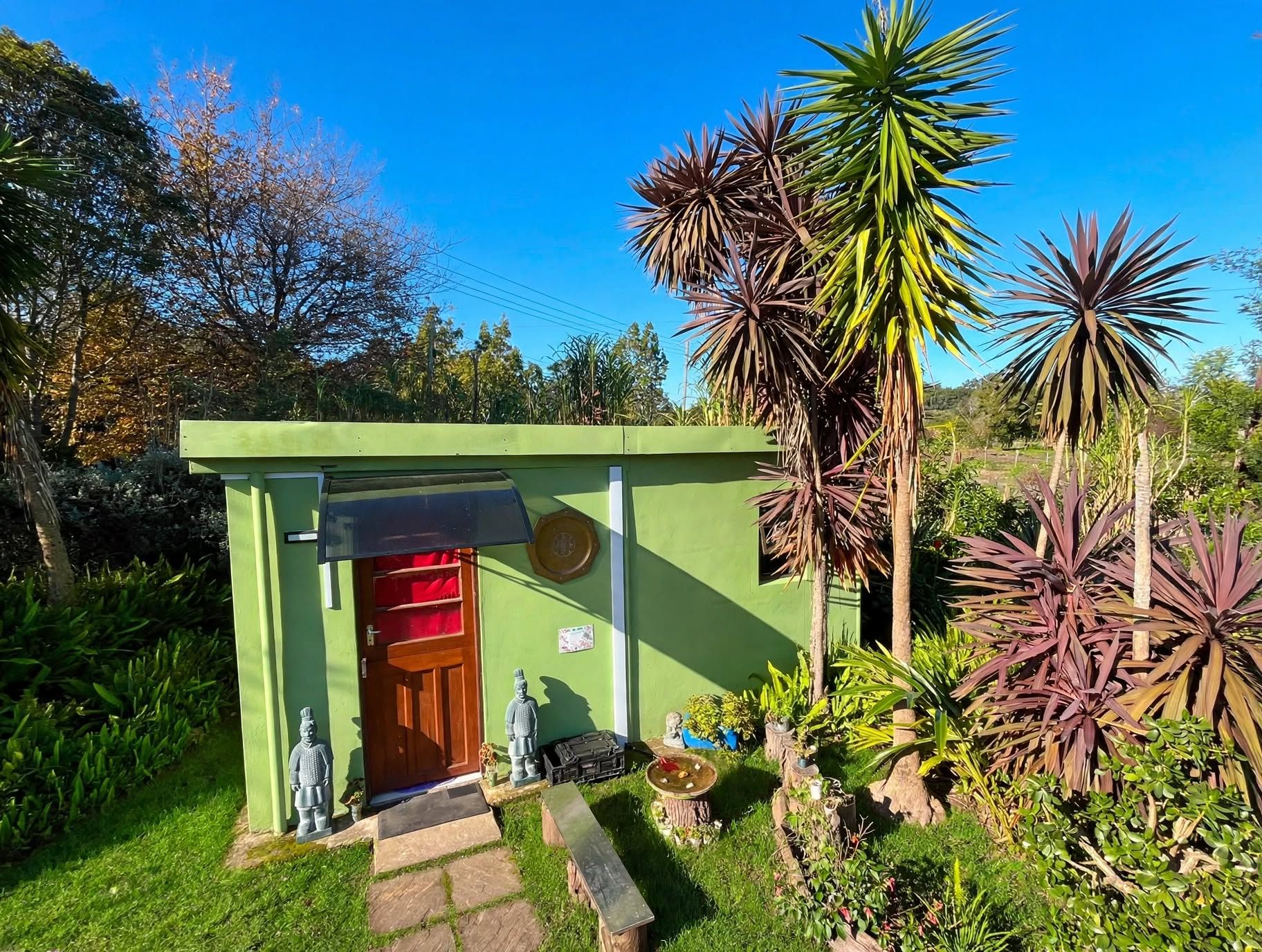 A small green structure with a red door, surrounded by various plants and a statue.