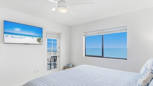 Quarto com cama, televisão, ventilador de teto, janela com vista para o oceano e porta que dá para uma varanda.