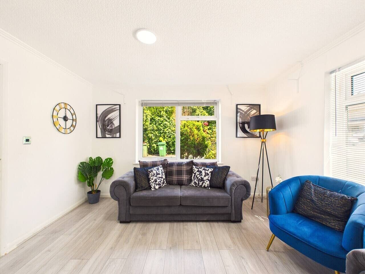 En stue med en grå sofa, en blå lænestol, en gulvlampe og et vindue med udsigt til grønne omgivelser.