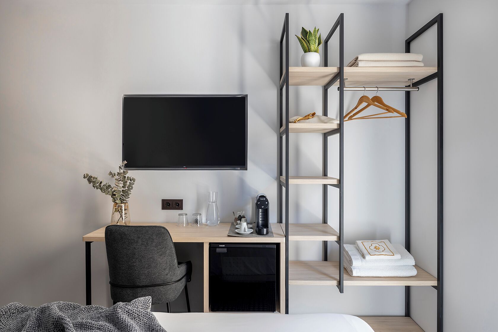 A compact hotel room with a desk, chair, bed, TV, shelves, and a potted plant.