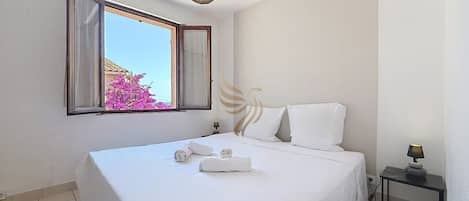 A hotel room with a bed, a window with curtains, a bedside table with a lamp, and a wall-mounted light fixture.