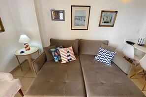 A modern living room with a brown sofa, two framed pictures on the wall, a small side table with a lamp, and a white chair. - SHB Piscina vista mar e pé na areia (Santos)