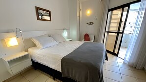 A bedroom with a bed, bedside tables, a nightstand, a wall-mounted light, a window with curtains, and a view of buildings outside. - SHB Piscina vista mar e pé na areia (Santos)