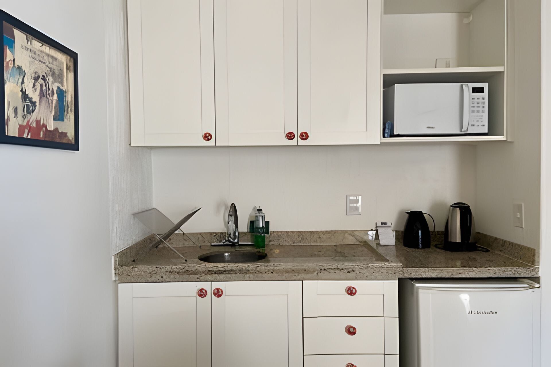 Una cocina compacta con armarios blancos, microondas, hervidor y refrigerador.