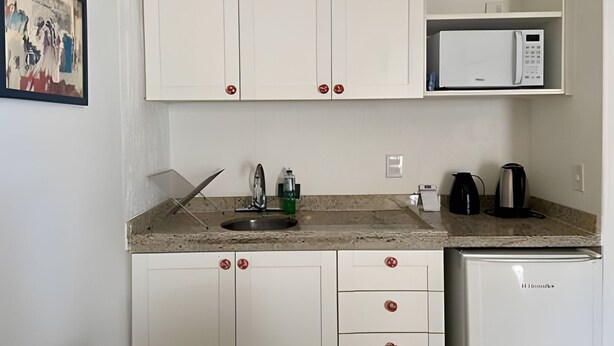A compact kitchen with white cabinets, a microwave, a kettle, and a refrigerator. - SHB Piscina vista mar e pé na areia (Santos)