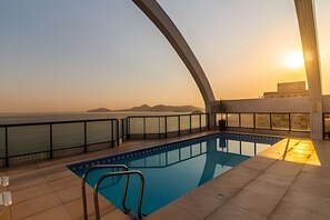 A rooftop swimming pool with a view of the ocean and distant mountains during sunset. - SHB Piscina vista mar e pé na areia (Santos)