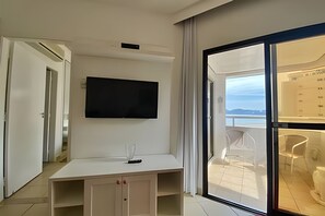 A modern hotel room with a flat-screen TV mounted on the wall, a small table with a glass, and a sliding glass door leading to a balcony with a view of the sea. - SHB Piscina vista mar e pé na areia (Santos)