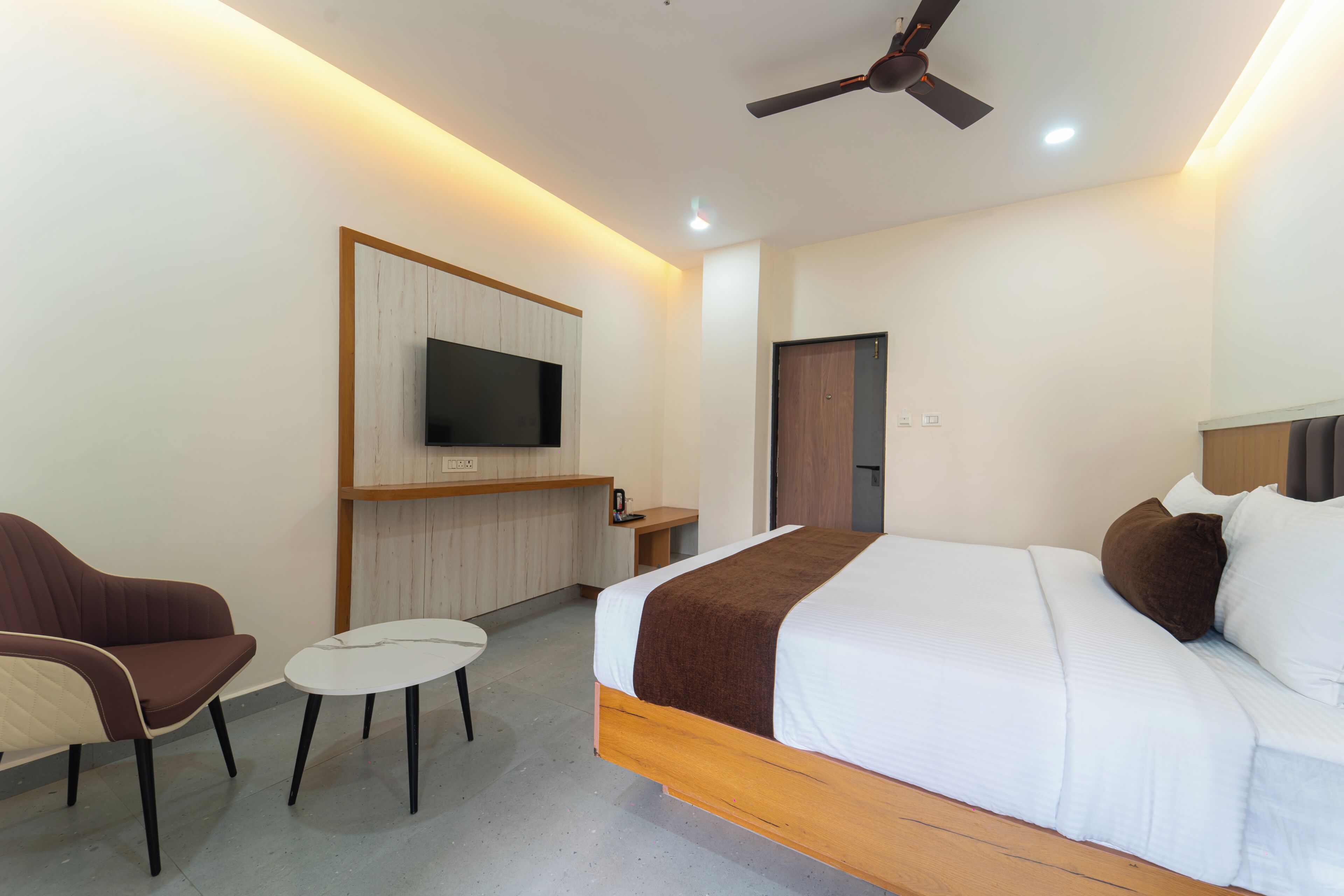 A hotel room with a bed, a TV mounted on a wooden panel, a ceiling fan, and a small seating area with a chair and table.