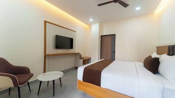 A hotel room with a bed, a TV mounted on a wooden panel, a ceiling fan, and a small seating area with a chair and table.