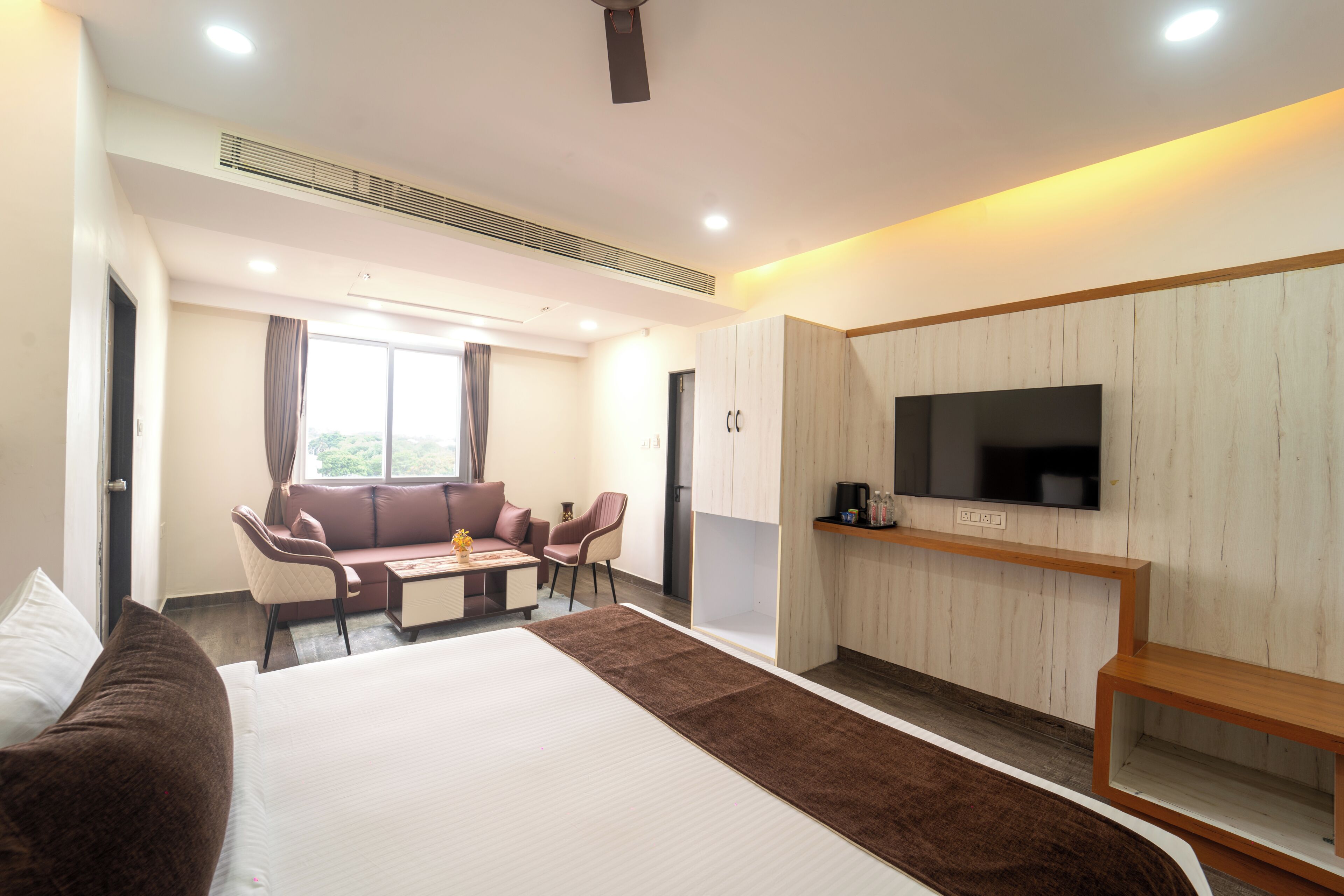 A modern hotel room with a large bed, a seating area with a sofa and chairs, a flat-screen TV mounted on the wall, and a small table with a coffee maker.