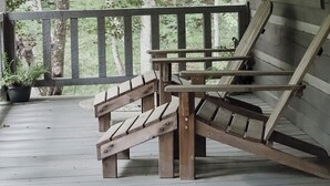 Een houten terras met een bank en een stoel, een hangende plantenbak en uitzicht op bomen.
