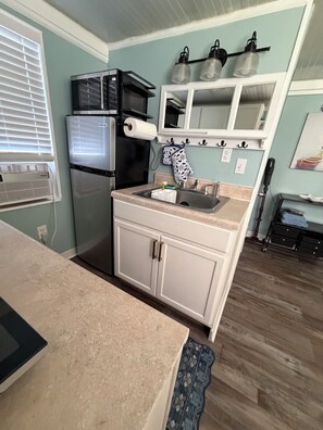 A compact kitchen with a refrigerator, stove, and sink. There is a window with blinds, a wall-mounted light fixture, and a patterned towel hanging on hooks. - Peaceful room with kitchenette in peaceful Homosassa w/boat parking on premises. (Homosassa)