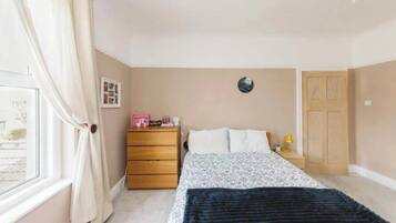 A bedroom with a bed, a dresser, a nightstand, a lamp, a window with curtains, and a door.