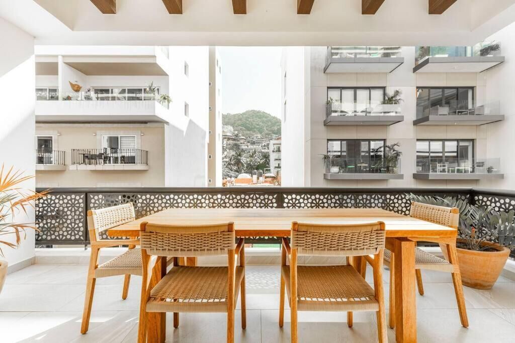 Un espace de restauration extérieur moderne, doté d’une table en bois et de chaises en osier, offrant une vue sur un paysage urbain avec des balcons.