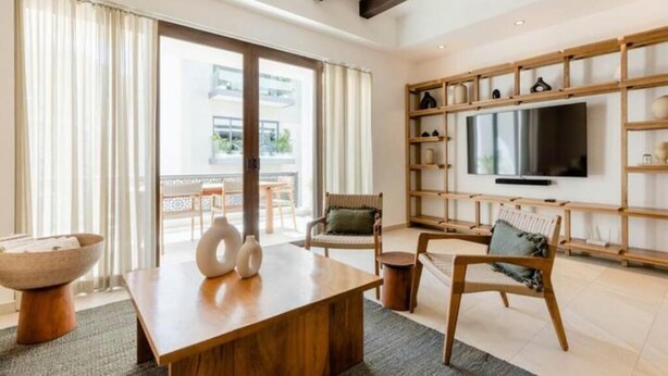 A modern living room with a wooden coffee table, two chairs, a television on a wooden shelf, and a large window with curtains. - Urban by Pardela (Puerto Vallarta)
