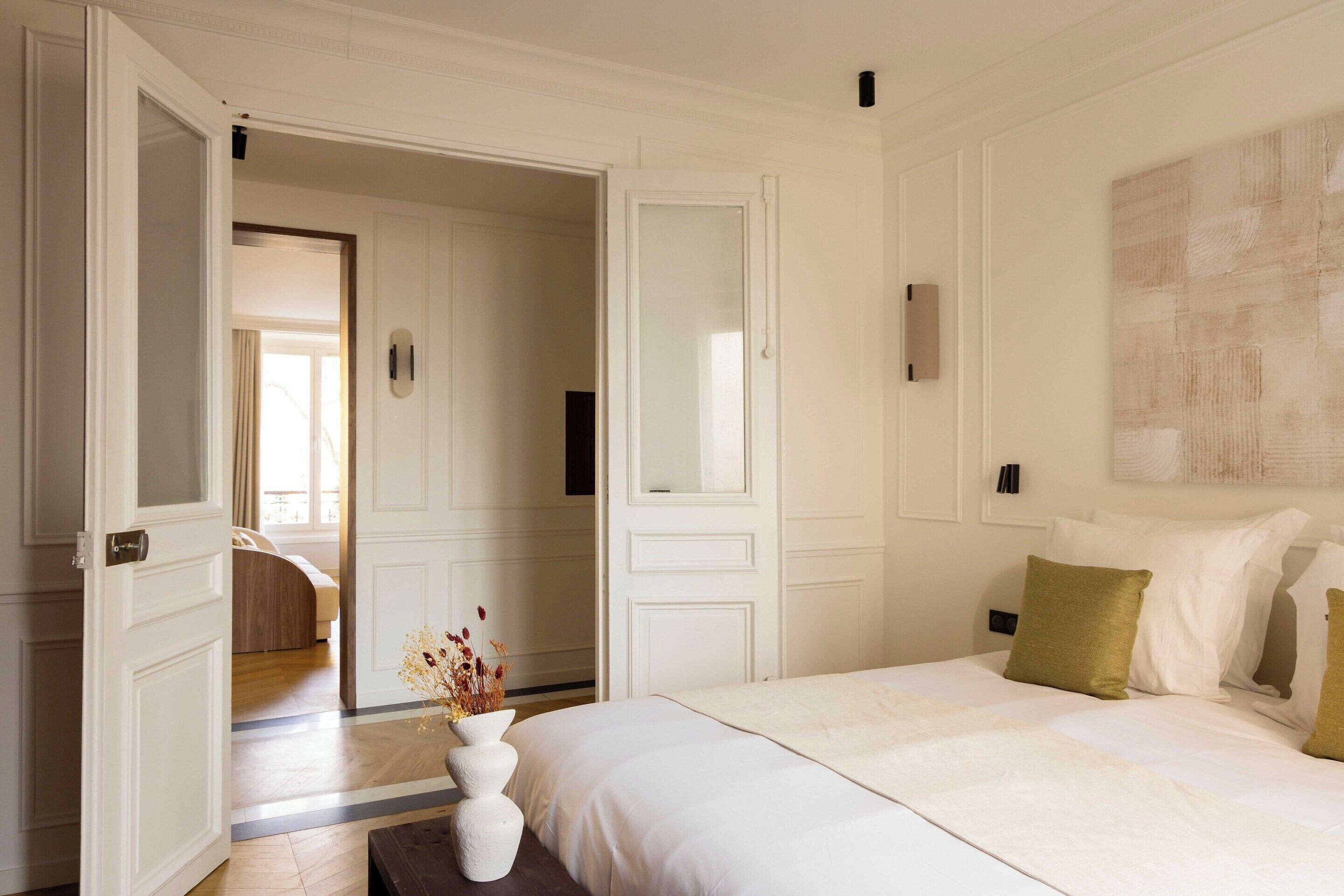 A hotel room with a bed, a white vase with flowers, and a large painting on the wall.