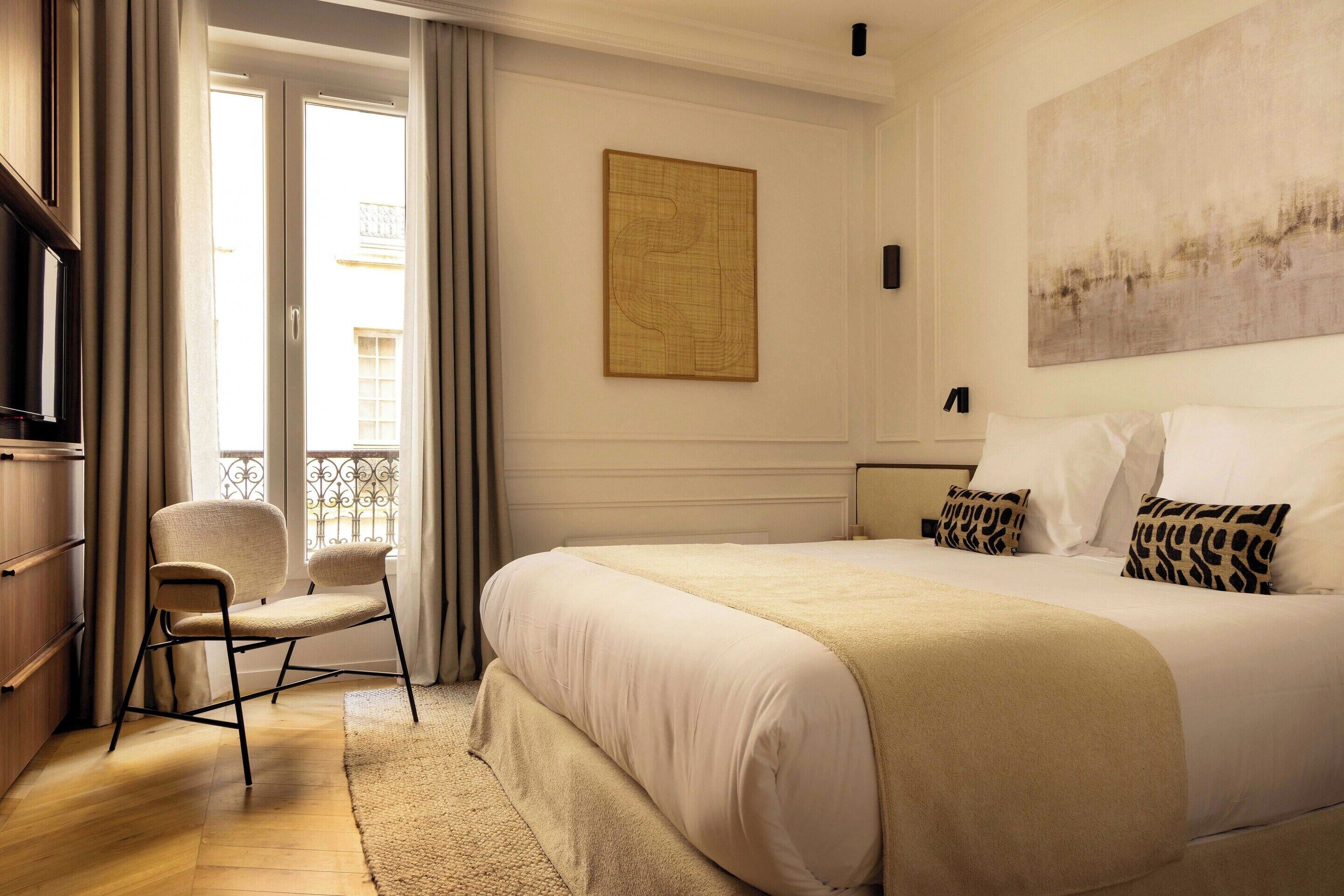 A hotel room with a bed, a chair, a TV, and a painting on the wall.