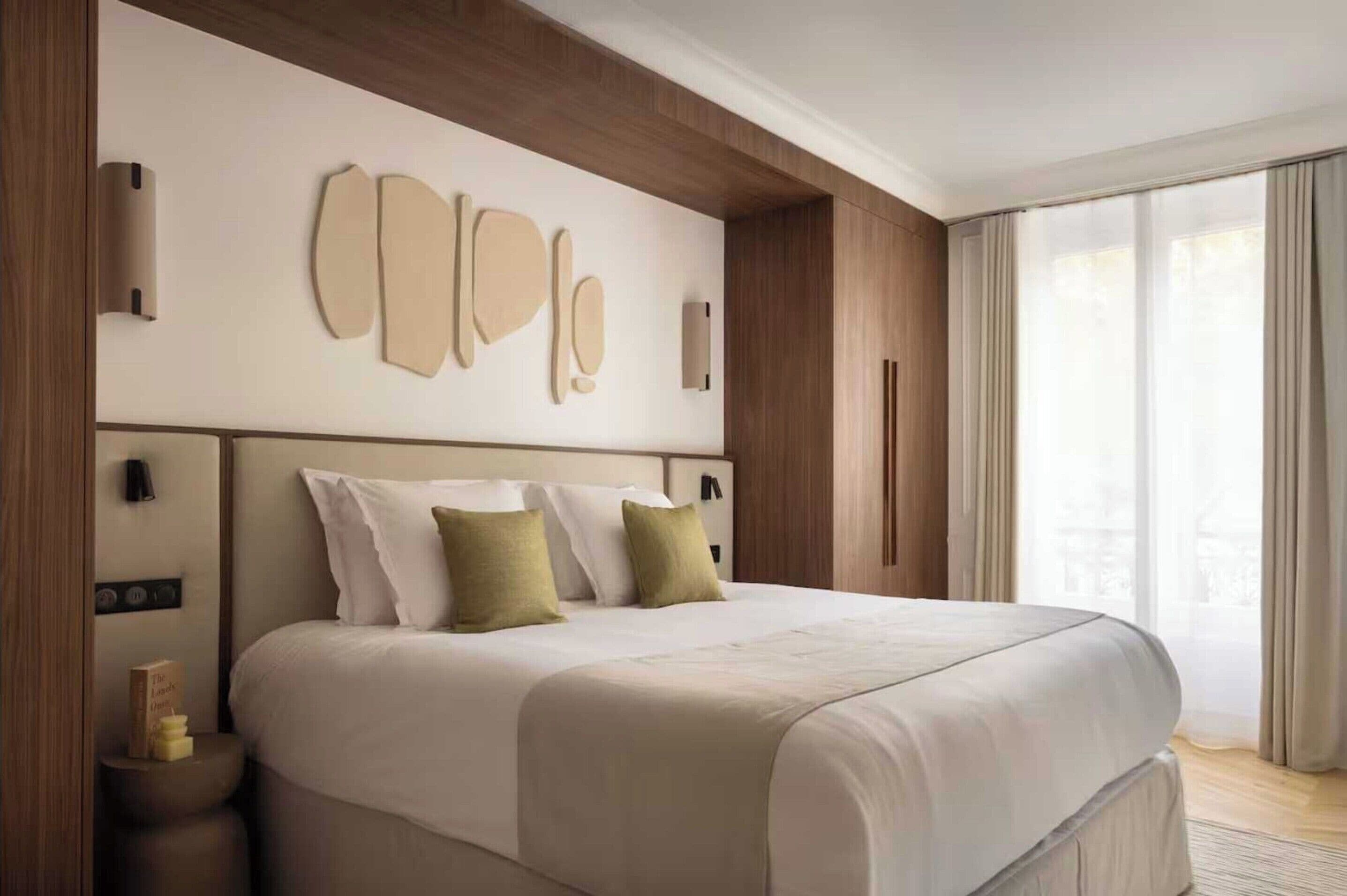 A hotel room with a large bed, a wooden headboard, a wall-mounted light fixture, and a large window with curtains.