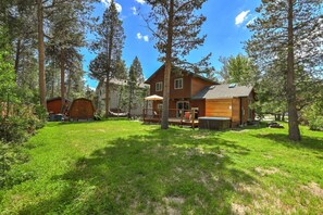 A two-story log cabin with a deck, surrounded by a grassy yard and tall pine trees. - The Rabbit Hole - Sauna. Soak. Repeat. (South Lake Tahoe)