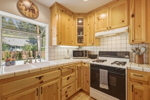 A kitchen with wooden cabinets, a stove, a microwave, and a window with a view of greenery. - The Rabbit Hole - Sauna. Soak. Repeat. (South Lake Tahoe)
