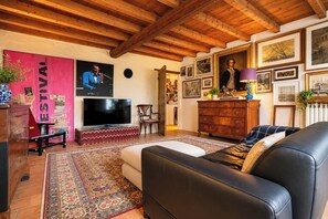 A living room with a TV, wooden ceiling, a sofa, a patterned rug, and a wall with framed pictures. - Le Stanze degli Artisti by Case al Lago (Monzambano)