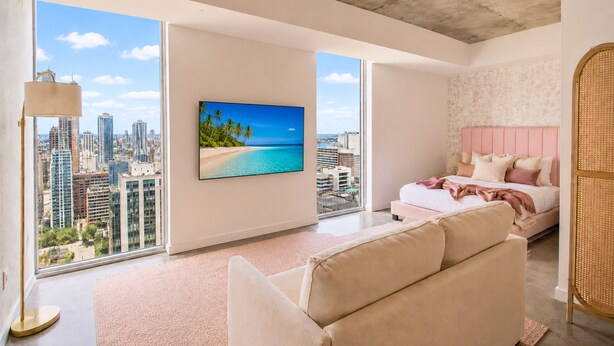 Ein modernes Hotelzimmer mit großem Fenster, einem Bett mit rosa Bettwäsche, einem beigefarbenen Sofa und Blick auf die Stadtlandschaft.