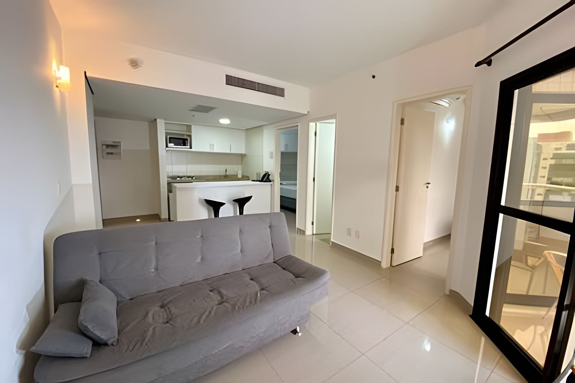 Una sala de estar moderna con un sofá gris, una cocina al fondo y un ventanal grande.