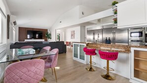 Una cuina moderna amb electrodomèstics d'acer inoxidable, taula de dinar de vidre amb cadires roses i zona de seients amb sofà i televisió.