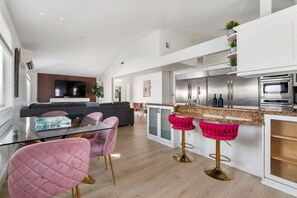 A modern kitchen with stainless steel appliances, a glass dining table with pink chairs, and a living area with a sofa and TV. - Family Pool House Oasis (National City)