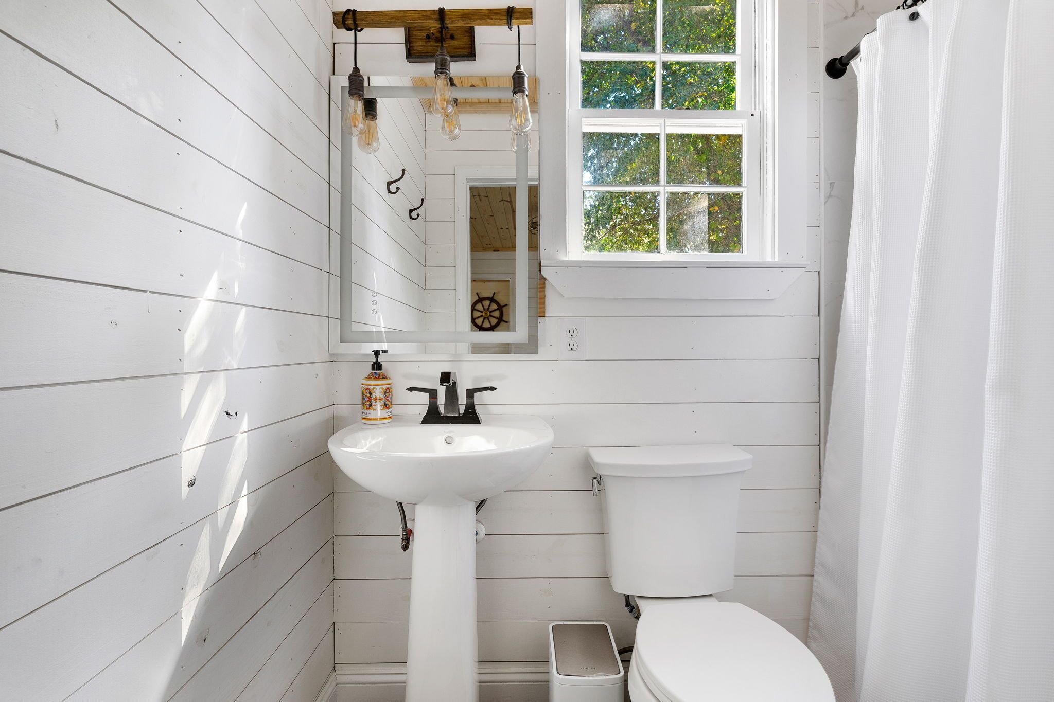 Une salle de bain avec des murs en bois peints en blanc, un lavabo blanc, des toilettes et une fenêtre donnant sur des arbres.
