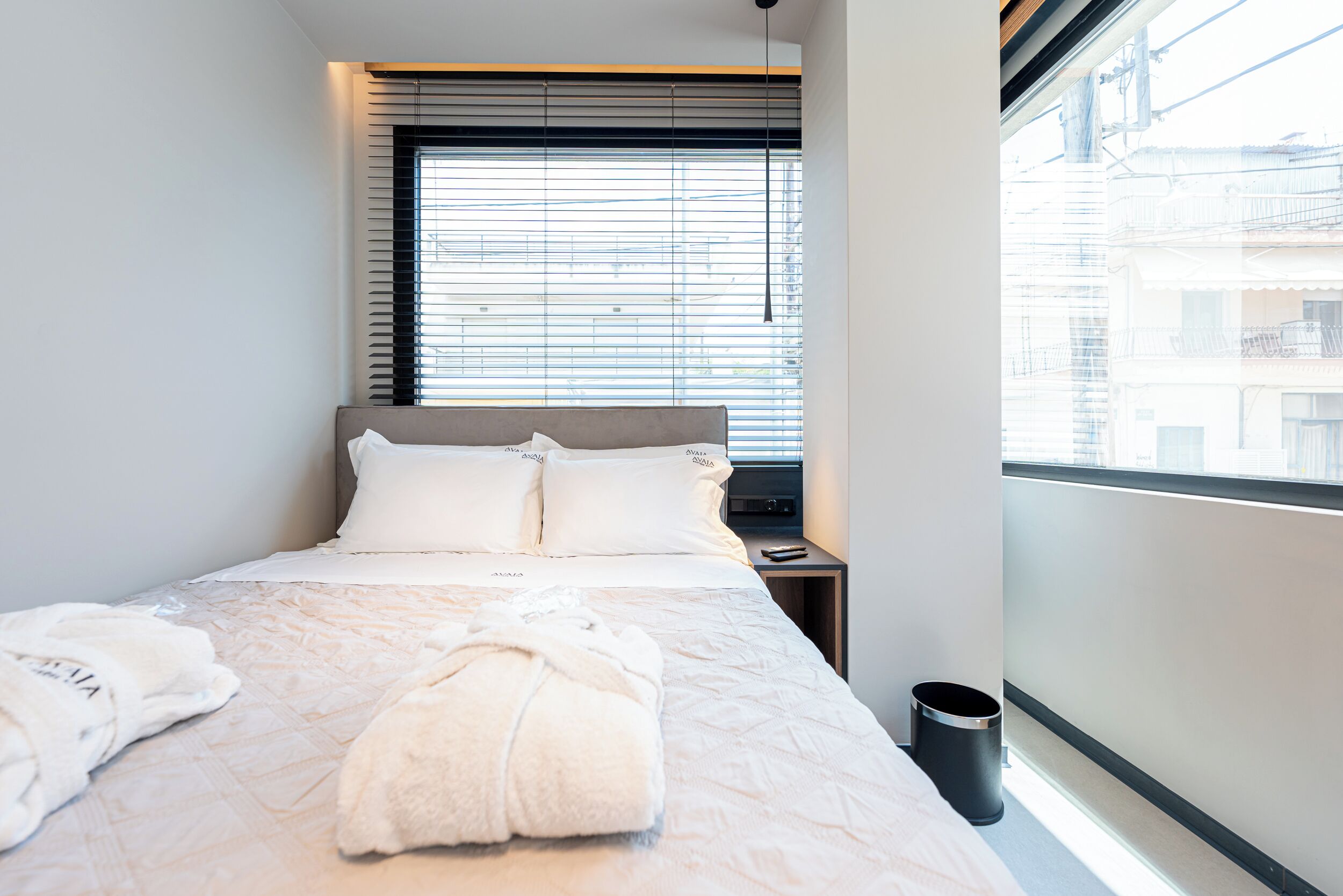 Una camera d'albergo con un letto, un comodino, una finestra con tende e vista sugli edifici.