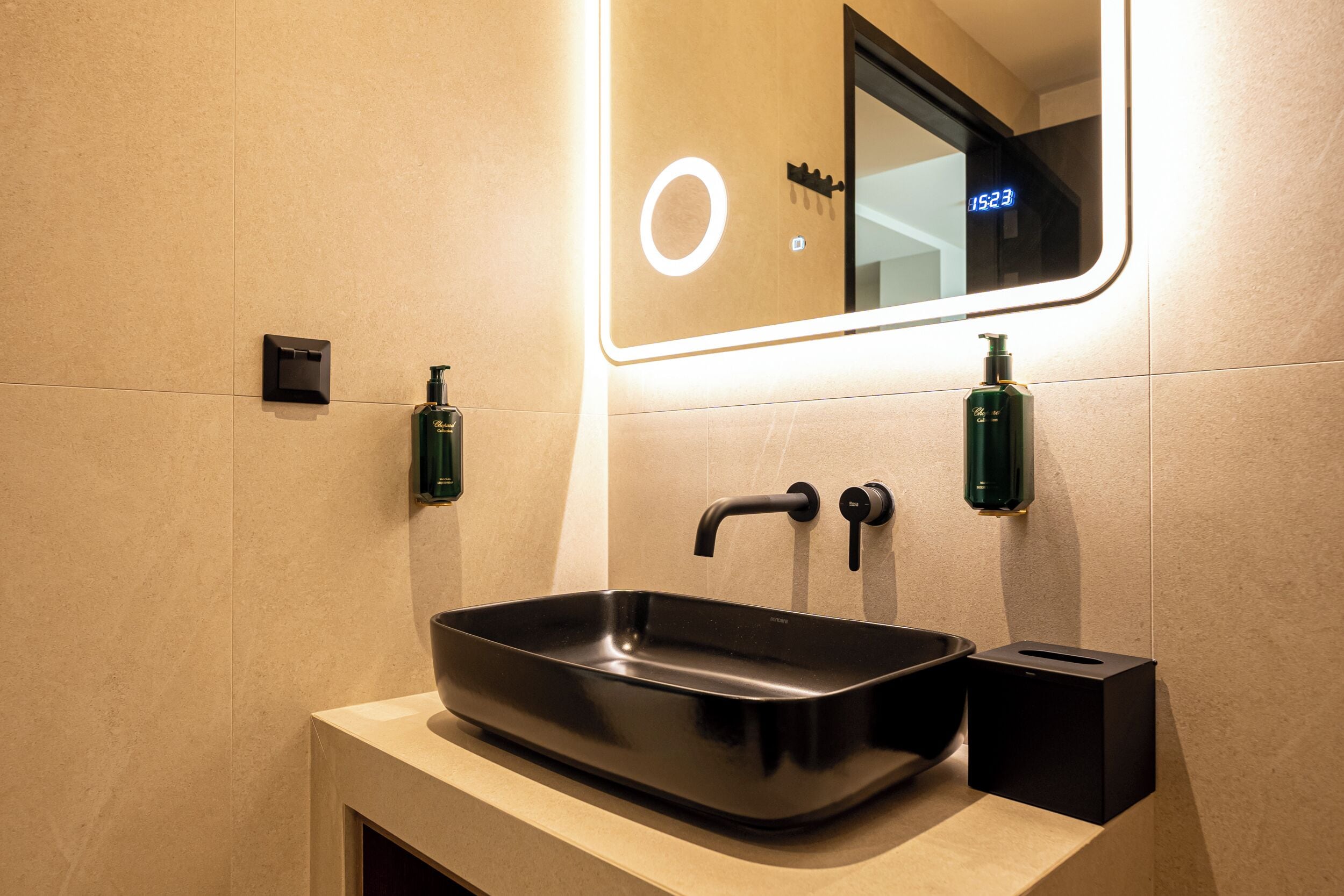 Un baño moderno con un lavabo rectangular, dos dispensadores de jabón, un espejo con luz y un cesto de basura.