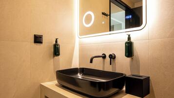 A modern bathroom with a rectangular sink, two soap dispensers, a mirror with a light, and a trash bin.