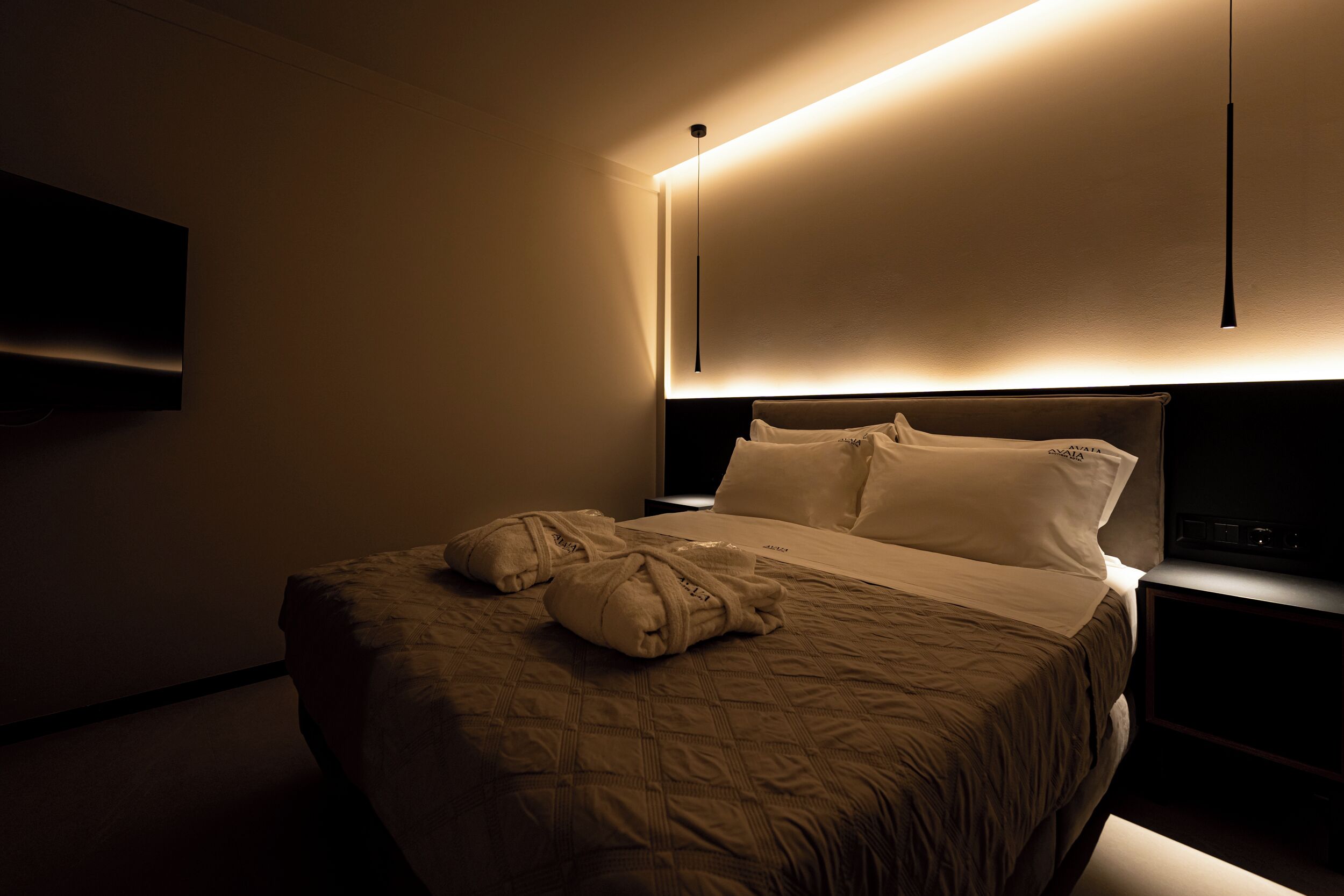 A hotel room with a large bed, a flat-screen TV mounted on the wall, and a bedside table with a lamp.