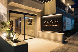 Entrada moderna de hotel com porta de vidro, uma placa indicando 'AVAIA Boutique Hotel' e um vaso com plantas.